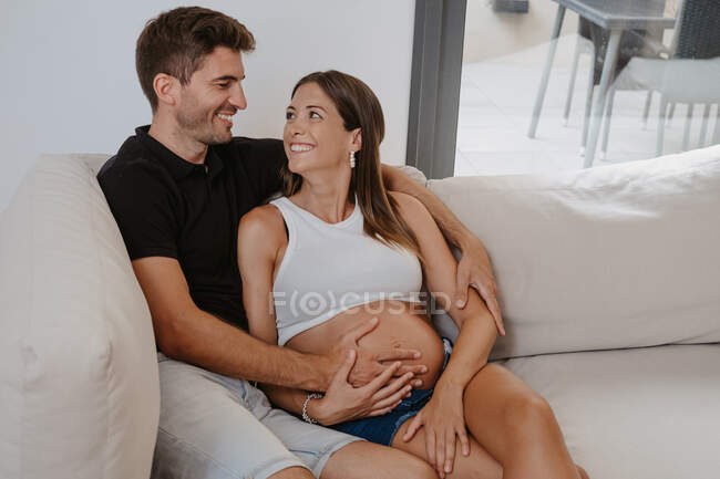 Homem abraçando barriga de expectante fêmea amada enquanto descansa no sofá na sala de estar olhando um para o outro — Fotografia de Stock