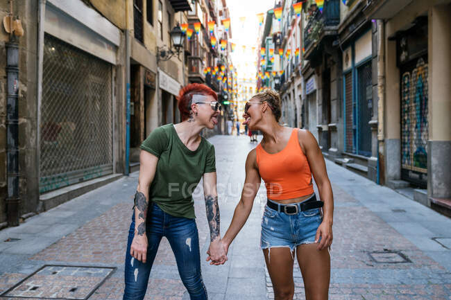 Cool jóvenes mujeres homosexuales con tatuajes en gafas de sol mirándose mientras sacan la lengua mirando de la mano en la pasarela de la ciudad - foto de stock
