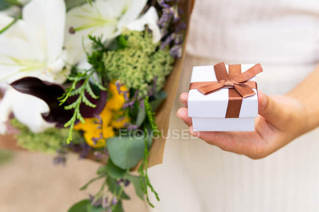 Ernte anonyme Frau mit kleinem Geschenkkarton und blühendem Blumenstrauß tagsüber auf verschwommenem Hintergrund — Stockfoto