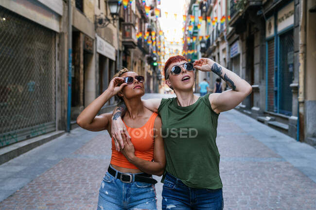 Coole junge homosexuelle Frauen mit Tätowierungen in Sonnenbrillen, die nach oben schauen, während sie sich auf dem Gehweg in der Stadt umarmen — Stockfoto