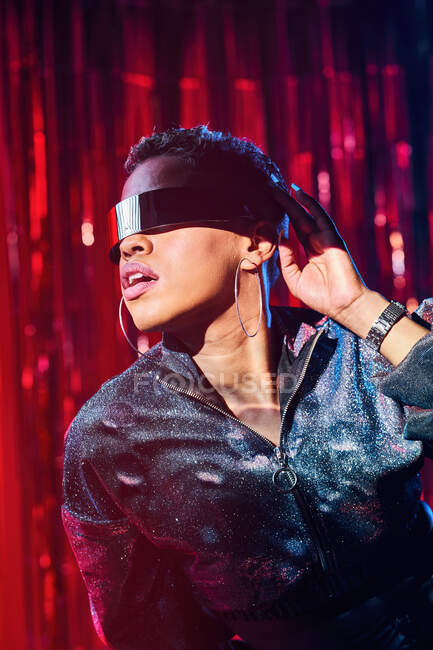 Trendy giovane femmina nera in occhiali cyberpunk con ombre sul viso in fasci di luce in discoteca — Foto stock
