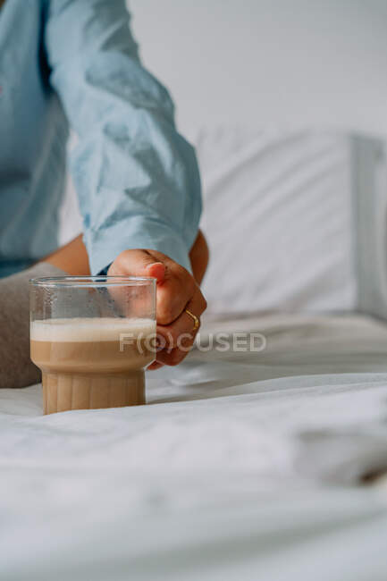 Coltivazione femminile irriconoscibile in anello con tazza di delizioso caffè con schiuma di latte in camera da letto — Foto stock