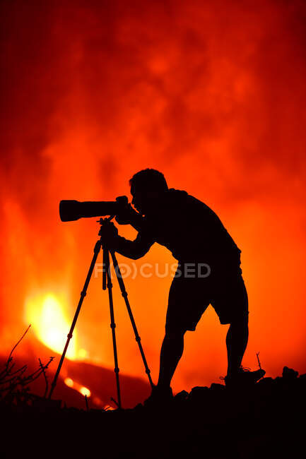 Vista lateral de la silueta de un hombre agachado fotografiando con un teleobjetivo y trípode la explosión de lava en La Palma Islas Canarias 2021 - foto de stock