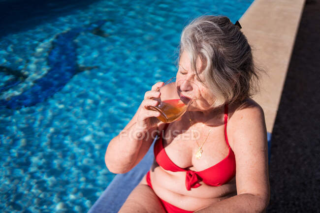 Высокий угол восторженной пожилой туристки в бикини, ярко смеющейся, расслабляясь у бассейна с напитком — стоковое фото