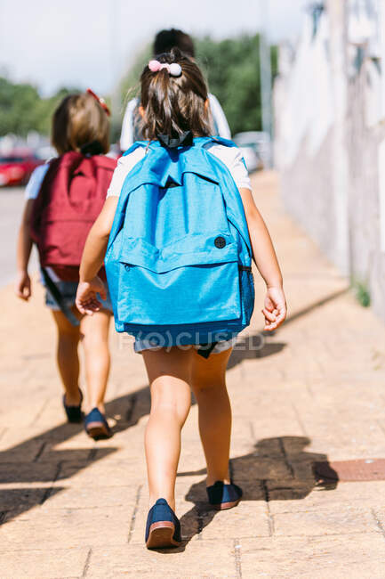 Back view of faceless schoolkids with rucksacks strolling on tiled pavement in city on sunny day — Stock Photo