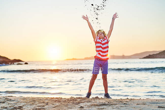 Повне тіло безтурботного хлопчика в літньому одязі кидає пісок на узбережжя махаючого моря на заході сонця — стокове фото