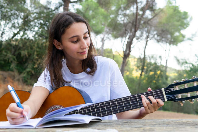 Adolescente femenina con guitarra clásica tocando el acorde mientras escribe música en copybook en el parque sobre fondo borroso - foto de stock