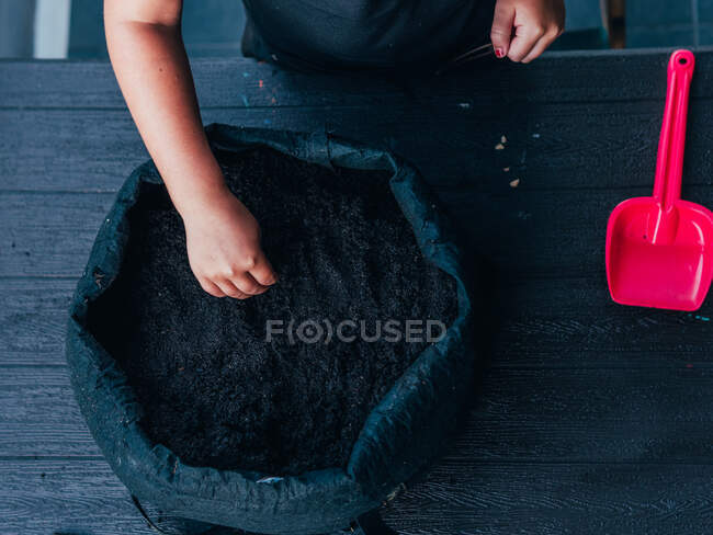 From above of crop anonymous child standing at dark table and touching dry black soil in pot in daytime — Stock Photo