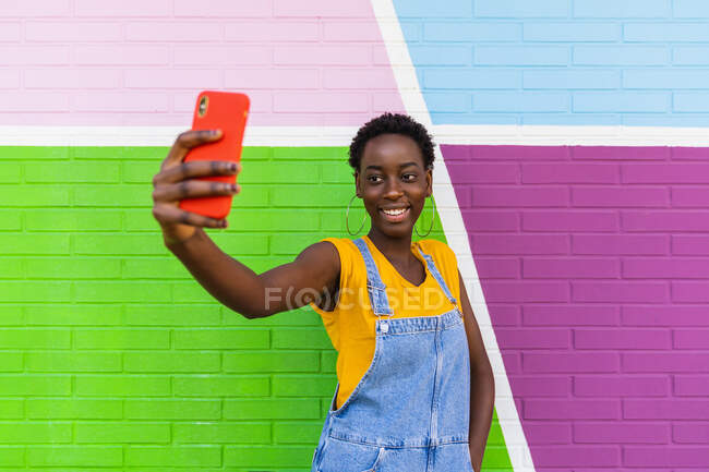 El foco selectivo del teléfono celular en las manos de la mujer alegre afroamericana tomando el autorretrato contra la pared brillante - foto de stock