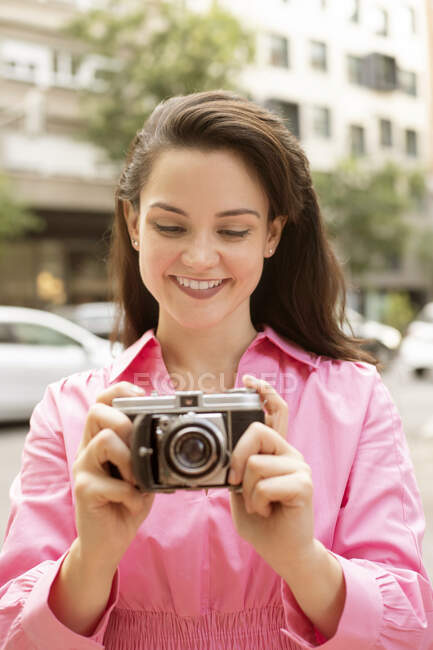 Young happy female with long brown hair taking picture on old fashioned photo camera on street in city — Stock Photo