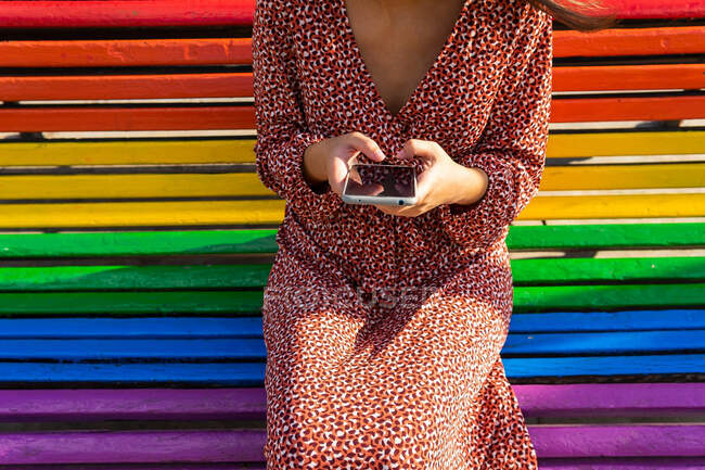 Обрізати невпізнавану жінку в одязі, сидячи на барвистій лавці і переглядаючи мобільний телефон вдень — стокове фото