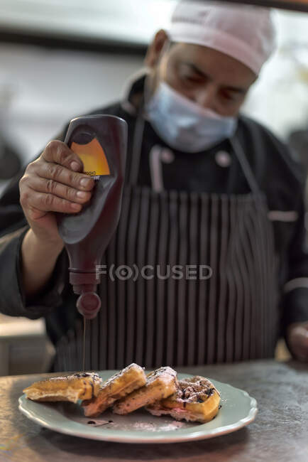 Crop cocinero masculino étnico en máscara estéril decorando deliciosos gofres vieneses con salsa de chocolate de la botella en la cocina del restaurante - foto de stock