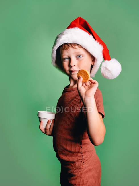 Vista lateral del adorable niño con el sombrero de Navidad de Santa tomar la galleta de la taza contra el fondo verde mirando a la cámara - foto de stock