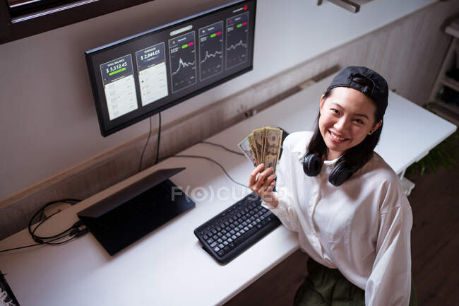 From above of cheerful young ethnic female dealer cooling off with banknotes while looking at camera against monitor with diagrams — Stock Photo
