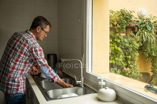 Vista lateral da calma pensativo maduro macho lavar pratos sujos enquanto está perto de pia na cozinha e fazer tarefas domésticas — Fotografia de Stock