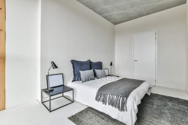 Kreative Gestaltung des Schlafzimmers mit Kissen auf Bettdecke auf dem Bett zwischen Tisch mit Lampe — Stockfoto