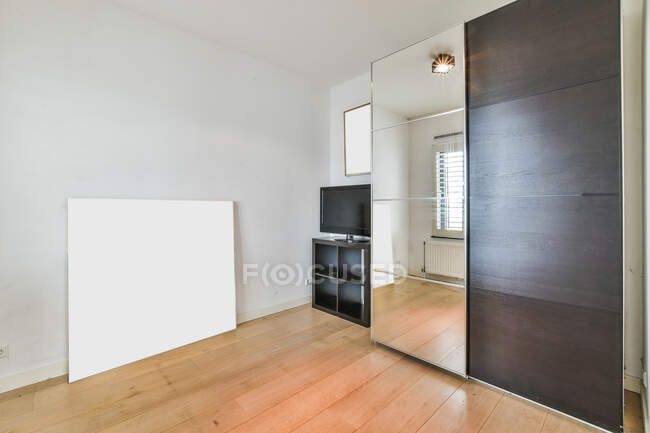 Habitación contemporánea con espejo en el armario contra la televisión con pantalla negra y panel en parquet en casa de luz - foto de stock