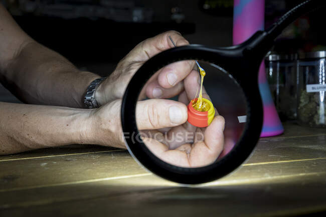 Crop unrecognizable male showing jar with marijuana glue and stick through magnifying lamp in workspace — Stock Photo