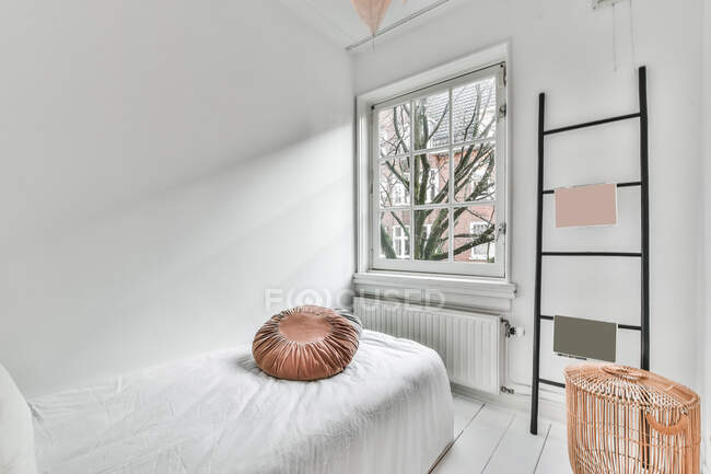 Удобная односпальная кровать с подушкой в спальне в стиле минимализма с лестницей и корзиной в современной квартире с большими окнами — стоковое фото