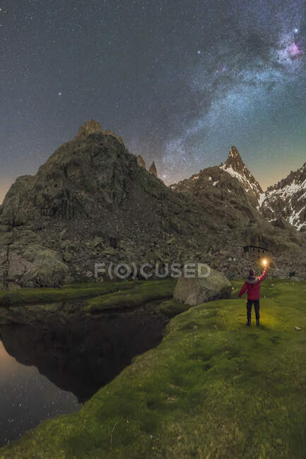 Back view of unrecognizable male tourist with torch admiring snowy mounts reflecting in water under starry sky at night — Stock Photo
