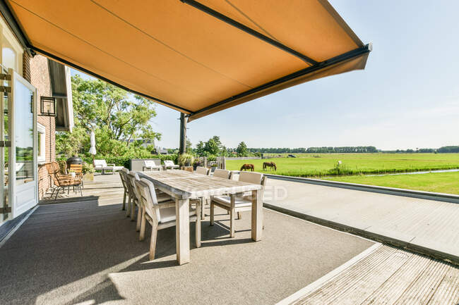 Table and armchairs under canopy on veranda against stallions feeding grass in countryside field under cloudy sky — Stock Photo