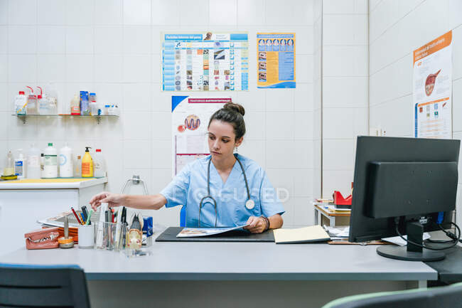 Увага молода жіночий ветеринар у формі з стетоскопом займає місце під час дивлячись на стіл з комп'ютером у лікарні. — стокове фото