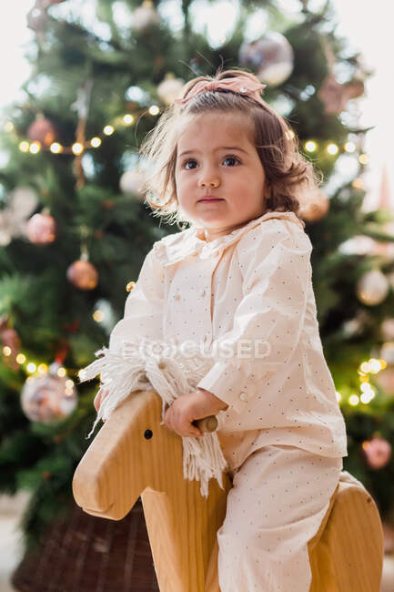 Adorable little toddler girl sitting on wooden rocking horse near Christmas tree decorated with fairy lights and toys — Stock Photo