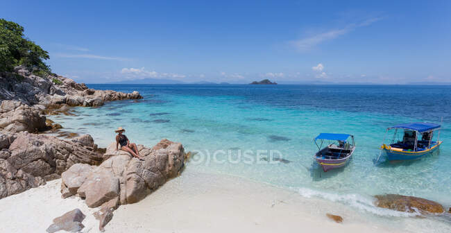 Viajera en sombrero sentada sobre piedras en la orilla arenosa del mar turquesa claro en Malasia - foto de stock