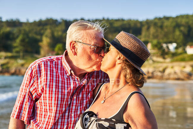 Gentle senior couple hugging and kissing each other on wet shore of pond in sunny day — Stock Photo