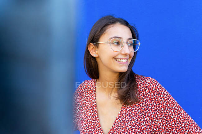 Mujer feliz en vestido rojo con estilo de pie contra la pared azul en el día mirando hacia otro lado - foto de stock