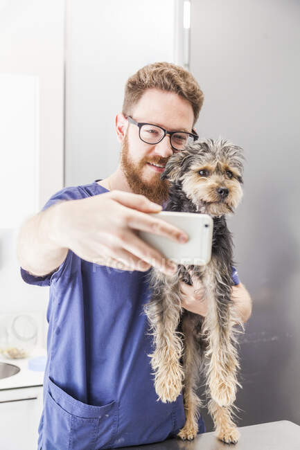 Médico veterinário masculino positivo tomando auto retrato com Yorkshire Terrier lambendo bochecha na clínica veterinária — Fotografia de Stock