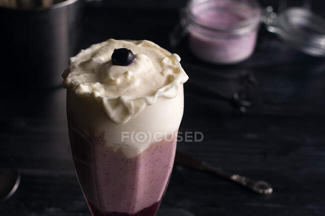 Glass of delicious banana and blueberry smoothie with whipped cream on chopping board against jar of yogurt and metal bucket — Stock Photo