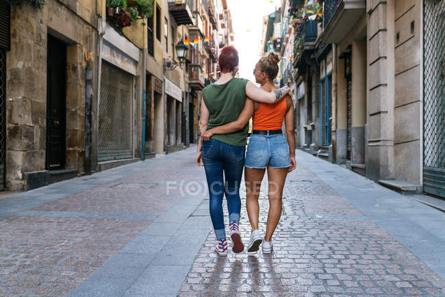 Vista posterior de mujeres homosexuales jóvenes irreconocibles con tatuajes en gafas de sol abrazándose mientras camina por la pasarela en la ciudad - foto de stock