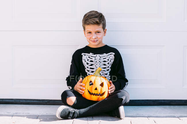 Ragazzo sorridente preadolescente che indossa costume nero di Halloween con stampa scheletro in piedi vicino scolpito Jack O Lanterna zucca contro muro bianco — Foto stock
