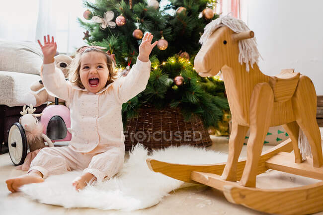 Full body of happy girl with raised arms sitting on rug near rocking horse and Christmas tree — Stock Photo