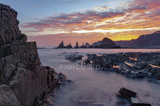Espectacular paisaje con olas de mar espumosas lavando formaciones rocosas rugosas de diversas formas en la playa salvaje de Geirua en Asturias España durante la puesta del sol - foto de stock