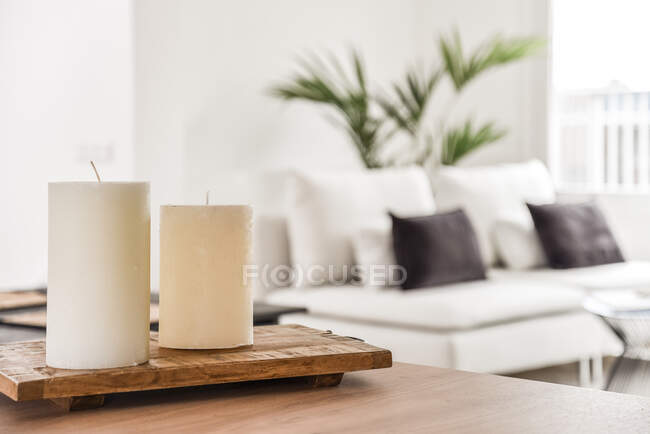 Velas brancas colocadas na mesa de madeira contra sofá confortável perto da planta envasada na sala de estar — Fotografia de Stock