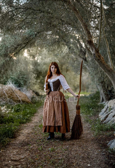 Bruxa séria de vestido de pé com livro mágico de feitiços e vassoura na estrada na floresta e olhando para a câmera — Fotografia de Stock