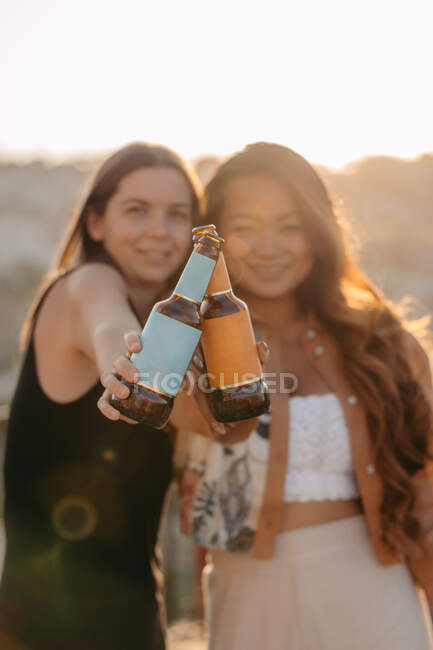 Positive junge multiethnische Freundinnen lächeln fröhlich und klimpern mit Bierflaschen, während sie bei Sonnenuntergang an der Terrassenbar in Kappadokien, Türkei, eine angenehme Zeit miteinander genießen. — Stockfoto