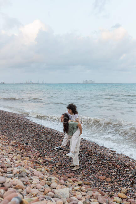 Allegro giovane femmina dando fidanzata lesbica cavalcata mentre si diverte sulla riva di ciottoli contro il mare ondulato al crepuscolo — Foto stock