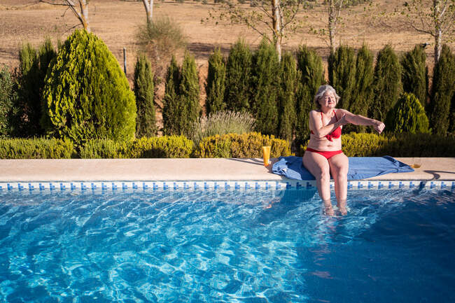 Старша жінка-туристка, що захищає шкіру сонцезахисним лосьйоном під час сонячних ванн на березі басейну під час літніх канікул — стокове фото