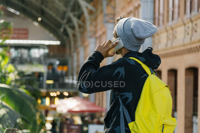 Вид ззаду анонімного чоловічого хіпстера з яскраво-жовтим рюкзаком, що стоїть біля ескалатора під час розмови на смартфоні — стокове фото