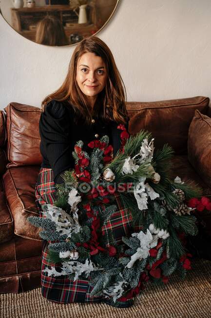 Donna allegra in abiti casual seduta con decorativa ghirlanda di Natale in mano sul divano e guardando la fotocamera in camera — Foto stock