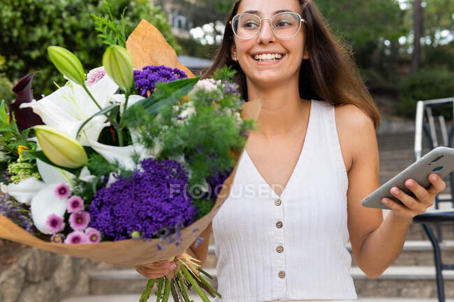 Contenu jeune femelle en lunettes avec bouquet de fleurs en fleurs messagerie texte sur téléphone portable dans les escaliers urbains — Photo de stock