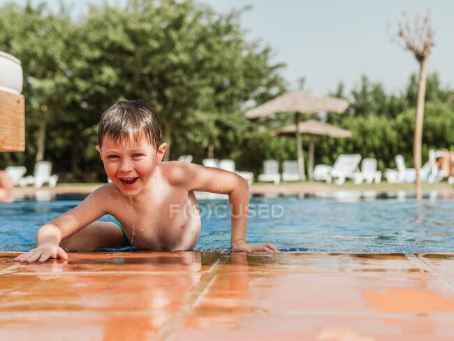 Criança bonita encantada com cabelo molhado inclinado na beira da piscina e olhando para a câmera enquanto se diverte durante o fim de semana de verão — Fotografia de Stock