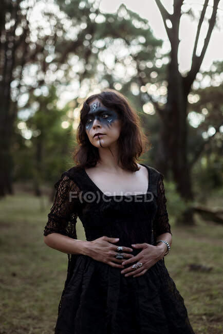 Mystic witch in long black dress and with painted face standing looking away in dark gloomy woods — Stock Photo