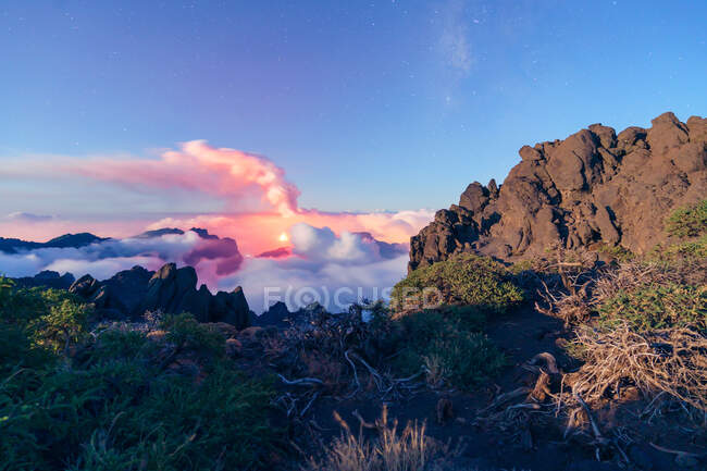 Night landscape with an erupting volcano in the background and a sea of clouds covering the mountains on a starry night from a vegetated and rocky mountain. Cumbre Vieja volcanic eruption in La Palma Canary Islands, Spain, 2021 — Stock Photo