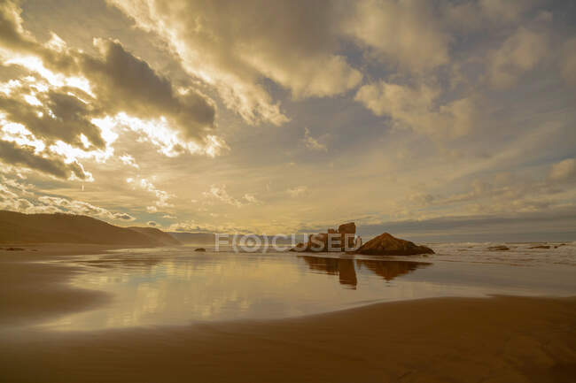 Scenic landscape of wet sandy shore with rocky formations under cloudy sky with sunlight in evening on beach Playon de Bayas in Asturais Spain — Stock Photo