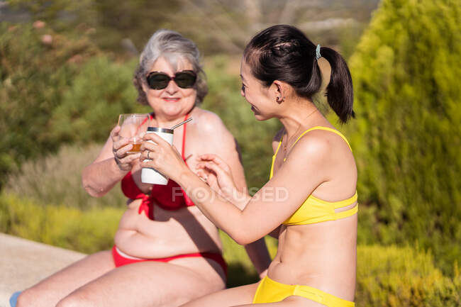Donna asiatica con coppa riutilizzabile seduta vicino alla vecchia con bicchiere di bevanda rinfrescante a bordo piscina — Foto stock