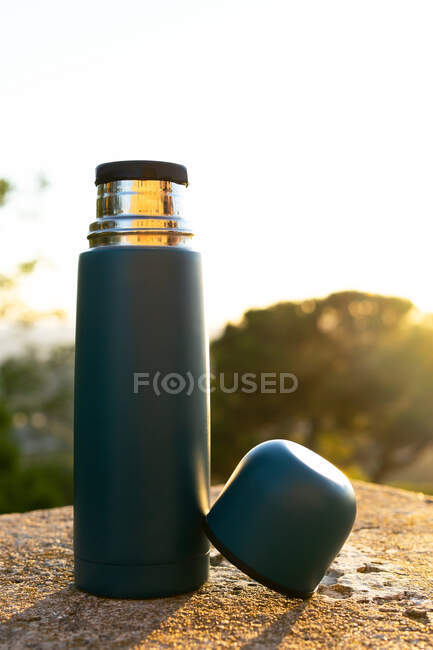 Modern stainless steel and plastic thermos with cup on rough surface under shiny sky at sundown — Stock Photo